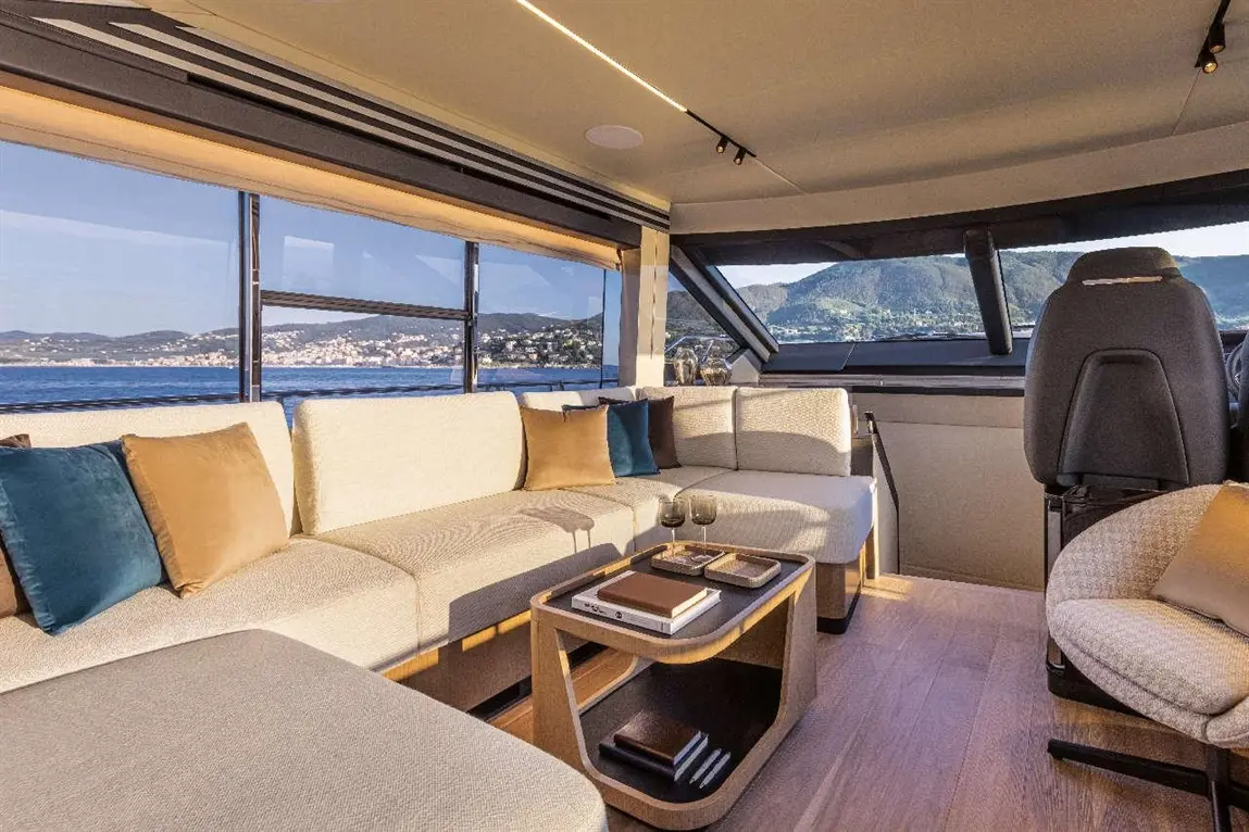 Luxurious yacht interior with beige sofas, decorative cushions, a coffee table, and panoramic views of mountains and sea.